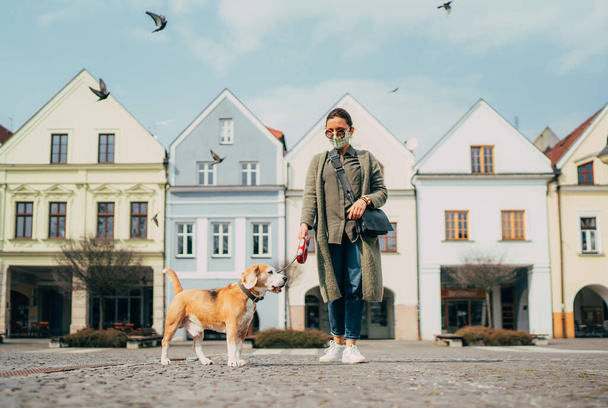 Young female fancy-dressed using a face mask as a coronavirus spreading prevention walking with her beagle dog on deserted old European squares and streets. Global COVID-19 pandemic concept image. - Photo, image