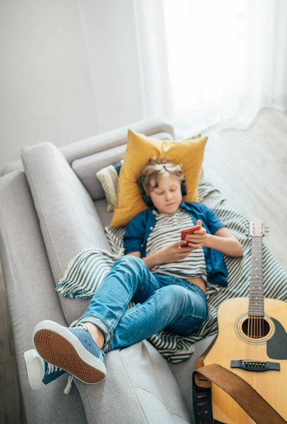 Preteen boy lying with guitar on cozy sofa dressed casual jeans and new sneakers listening to music and chatting using wireless headphones connected with a smartphone. Using electronic devices concept - Photo, Image