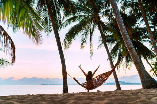 Young woman meeting morning sunrise sunlight sitting in a hammock and lazy stretching her body raising arms up on the sandy beach under the palm trees. Calm exotic places vacation concept image. - Fotoğraf, Görsel