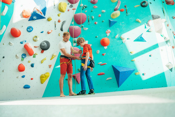 Father helping his teenage son and checking climbing harness near the indoor climbing wall. Boy preparin - Fotografie, Obrázek