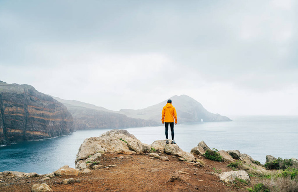 Trail runner man dressed orange waterproof jacket, running tights and shoes enjoying Atlantic ocean bay view on Ponta de Sao Loureno peninsula - the easternmost point of Madeira island, Portugal - Фото, зображення