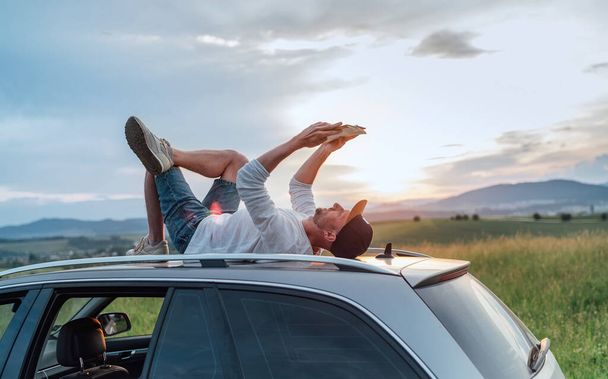 Middle-aged Man lying on the car roof and reading the paper bestseller book.He stopped his auto on the meadow with a beautiful valley view before the sunset. Reading a hobby or education concept image - Photo, Image