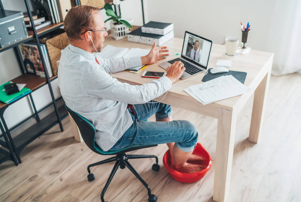  Comic modern home office situation. Businessman have a video call chatting on laptop and soaring his feet in Foot hot Bath under table. Distance work in worldwide quarantine time concept image - Fotoğraf, Görsel