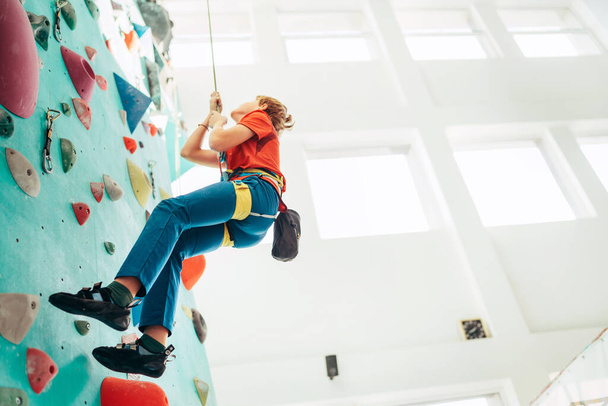 Teenager boy at indoor climbing wall hall. Boy is climbing using a top rope,chalk bag and climbing harness. Active teenager time spending concept image. - Foto, immagini