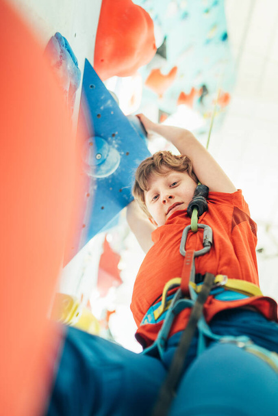 Teenager boy at indoor climbing wall hall. Boy is climbing using a top rope and climbing harness. He hanging on climbing holds and volume. Active teenager time spending concept image. - Photo, image