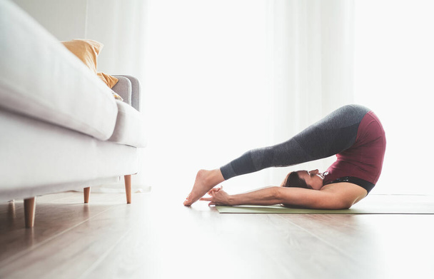 Young graceful Woman enjoying morning yoga exercises doing Halasana pose at home living room near the big window. Active people, body care and healthy lifestyle concept. - Fotografie, Obrázek