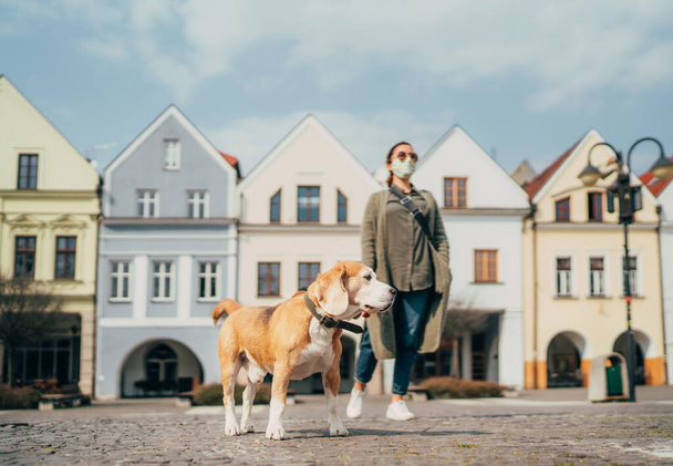 Young female fancy-dressed using a face mask as a coronavirus spreading prevention walking with her beagle dog on deserted old European squares and streets. Global COVID-19 pandemic concept image. - Фото, изображение