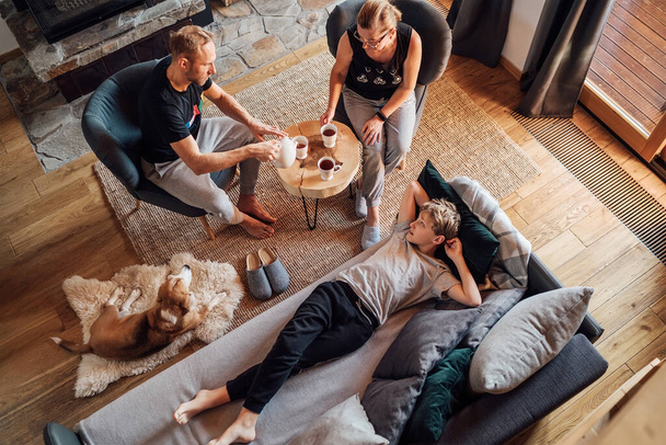 Peaceful family moments concept image. Cozy family drinking tea time. Father, mother and son at the home living room. Boy lying in comfortable sofa and stroking their beagle dog and smiling.  - Фото, изображение