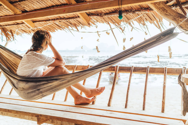 Young female sitting in hammock on straw hut terrace and enjoying a seaside beach landscape on Tonsai Beach in Thailand. Careless summertime exotic vacation concept image. - Foto, imagen