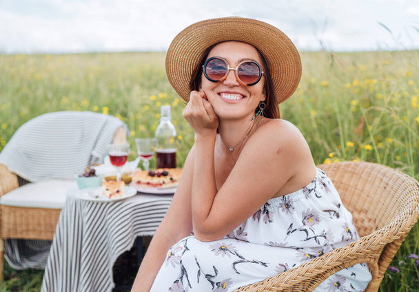 Sincerely smiling at camera young Woman in sunglasses and  straw hat dressed light summer dress sitting in rattan chair on the high green grass meadow near picnic table cheerful.Natural beauty concept - Zdjęcie, obraz