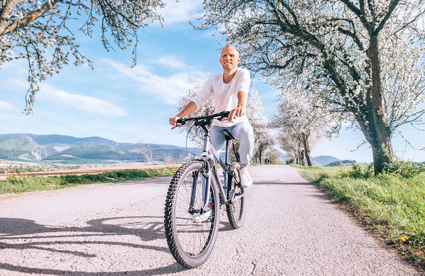 Happy smiling man riding a bicycle on the country road under blossom trees. Spring is comming concept image. - Foto, Imagen