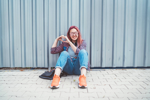Cheerful Beautiful modern smiling young female teenager in a checkered shirt and jeans with headphones sitting near a wall on the street and making a heart shape palms . Carefree student age concept. - Photo, Image