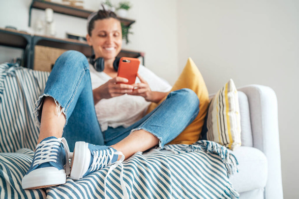 Young female sitting on cozy comfortable living room sofa, smiling and chatting with friends using modern smartphone. Selective focus on new blue sneakers. Comfortable clothing and footwear concept - Photo, Image