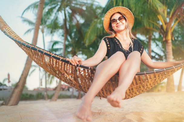 Happy smiling young woman in straw hat in sunglasses sitting in hammock and enjoy morning sun light on the Koh Samui tropical beach, Thailand. - Foto, Imagem