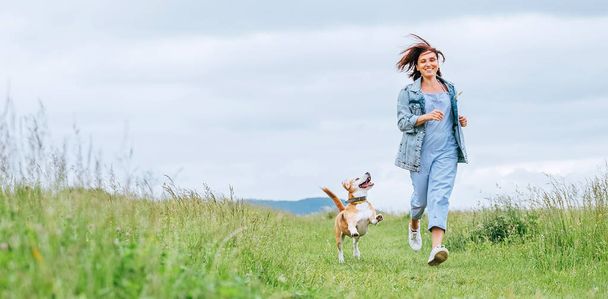 Happy smiling jogging female with fluttering hairs and her beagle dog running and looking at eyes. Walking by meadow grass path in nature with pets, healthy active people lifestyle concept image. - Photo, image