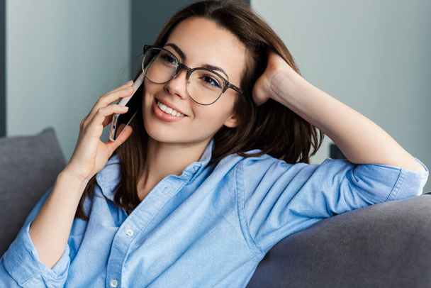 Image of cheerful woman in eyeglasses talking on cellphone and smiling while sitting at living room - Foto, Imagen