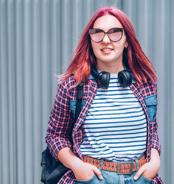 Beautiful modern smiling young female teenager with extraordinary hairstyle color in a checkered shirt and mirrored sunglasses portrait with headphones hanged on neck. Modern teens world concept image - Zdjęcie, obraz