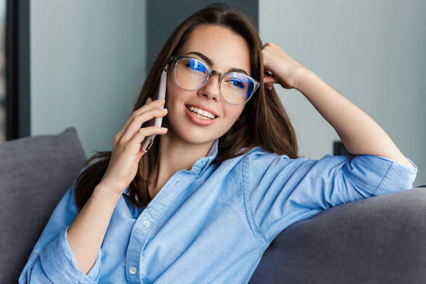 Image of cheerful woman in eyeglasses talking on cellphone and smiling while sitting at living room - Fotó, kép