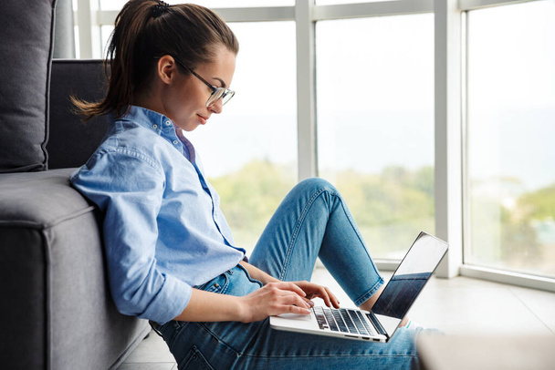 Obraz szczęśliwej kobiety w okularach pracującej z laptopem i uśmiechniętej siedzącej na podłodze w salonie - Zdjęcie, obraz