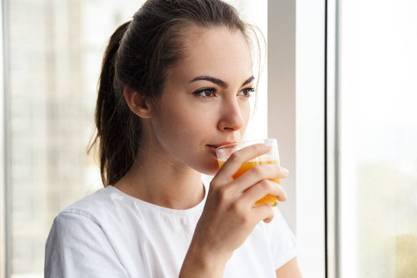 Image of focused young nice woman drinking fresh juice while looking at window indoors - Fotó, kép