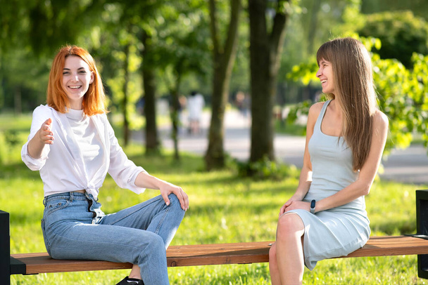 Two happy girls friends sitting on a bench outdoors in summer park chatting happily having fun. - Zdjęcie, obraz