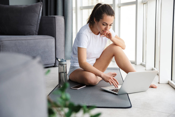 Image of brunette woman typing on notebook and cellphone while sitting on mat near window indoors - Foto, Imagen