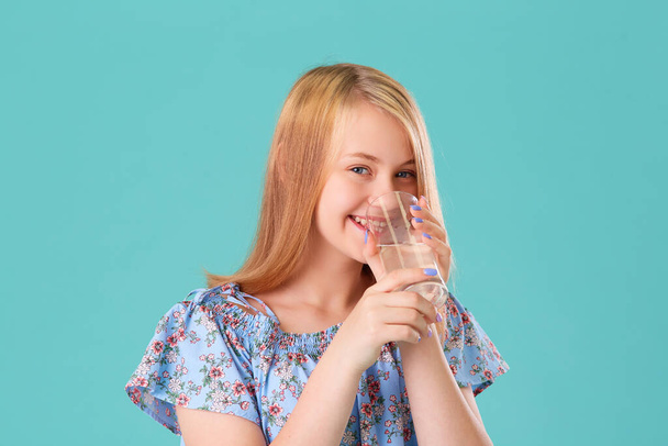 Nettes Teenie-Mädchen mit langen Haaren lächelt und trinkt ein Glas Wasser auf türkisfarbenem Hintergrund des Studios. - Foto, Bild