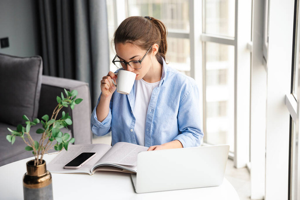 Image of beautiful focused woman reading book and drinking tea while working with laptop in living room - Foto, afbeelding
