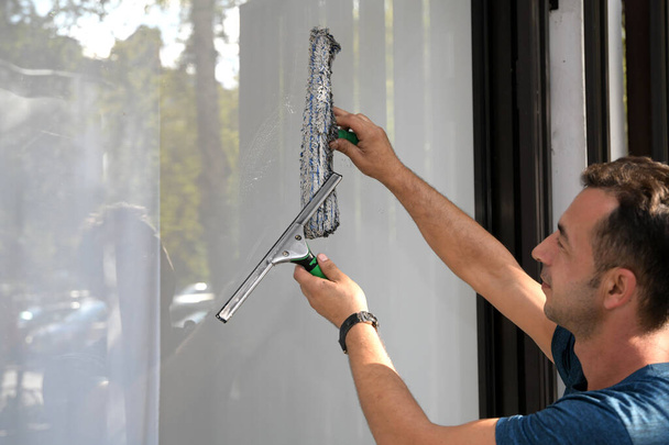 Καθαρίζω το παράθυρο. Ένας νεαρός άνδρας καθαρίζει και γυαλίζει τα παράθυρα με σφουγγάρι - Φωτογραφία, εικόνα