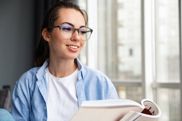 Image of cheerful caucasian cute woman in eyeglasses reading book and smiling while sitting in living room - Zdjęcie, obraz