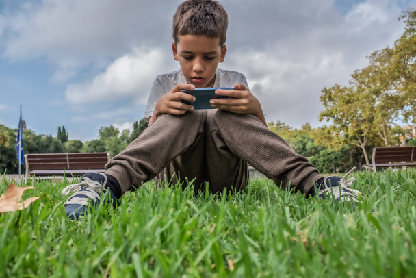 集中茶色の髪の少年は、スマートフォンでゲームをプレイ公園に座っている。子供の技術ガジェットへの中毒の概念 - 写真・画像