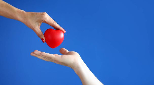 Женская рука передает красное сердце к мужской руке на синем фоне крупным планом. - Фото, изображение