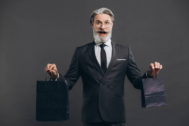 Business and fashionable bearded mature man in a gray suit with craft packages and making shopping on the grey background. - Foto, Bild