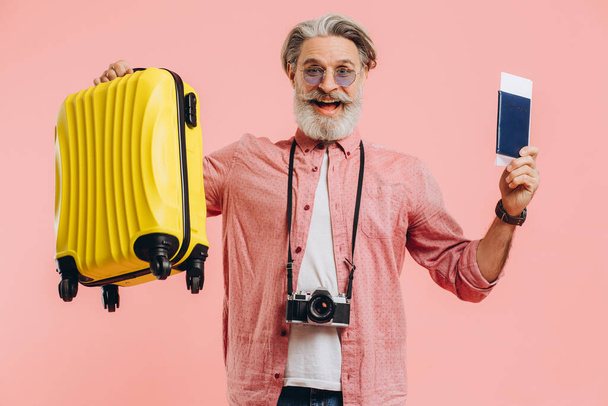 Κομψός γενειοφόρος άνδρας με φωτογραφική μηχανή κρατά κίτρινη βαλίτσα και διαβατήριο με εισιτήριο, χαμογελάει και προετοιμάζεται για το ταξίδι. - Φωτογραφία, εικόνα