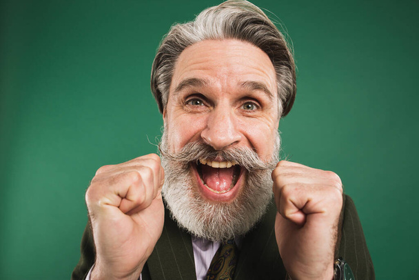 A middle-aged bearded man in a khaki suit shouts loudly and holds his fists against on a green background - 写真・画像
