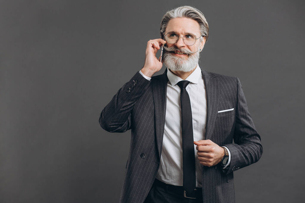 Business und modischer bärtiger älterer Mann im grauen Anzug lächelt und telefoniert vor grauem Hintergrund. - Foto, Bild