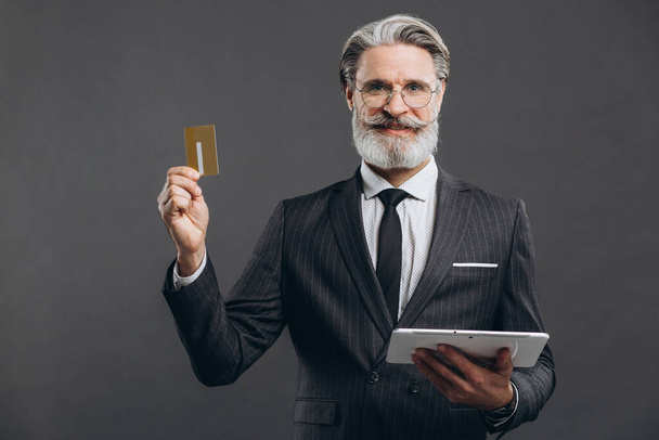 Homme mûr barbu d'affaires et à la mode en costume gris faisant du shopping en tablette avec sa carte de crédit en or sur le fond gris. - Photo, image