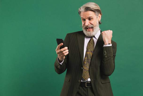 Sakallı, orta yaşlı, haki takım elbiseli bir adam yüksek sesle bağırıyor ve yeşil arka planda mutluluğun yumruğunu tutarken telefona bakıyor. - Fotoğraf, Görsel