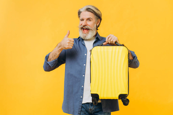 Стильный бородатый мужчина смеется, держа желтый чемодан и показывая крутой
 - Фото, изображение