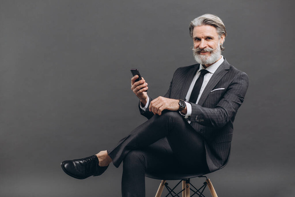 Деловой и модный бородатый мужчина зрелого возраста в спортивном костюме сидит и пользуется смартфоном на сером фоне
. - Фото, изображение