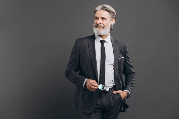 Obchodní a módní vousatý dospělý muž v šedém obleku drží brýle a usmívá se na kopírovací prostor na šedém pozadí. - Fotografie, Obrázek