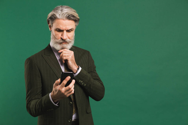 Stylish middle-aged man in khaki suit looks at phone and fixes mustache and beard on green background - Foto, Bild