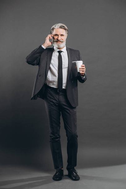 Деловой и модный бородатый мужчина в сером костюме разговаривает по телефону, пьет кофе и улыбается на сером фоне
. - Фото, изображение