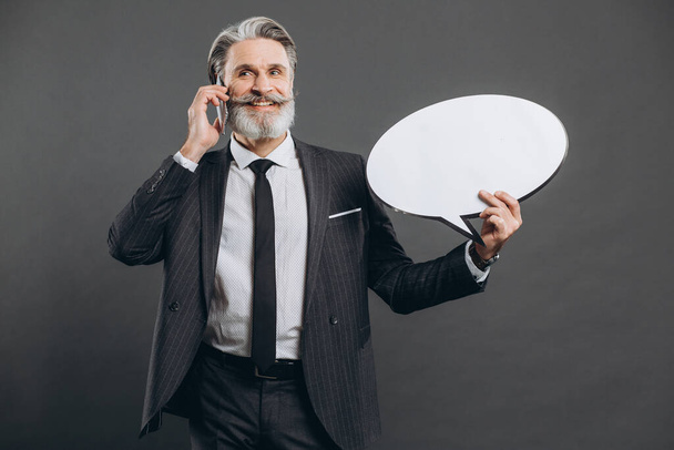 Obchodní a módní vousatý dospělý muž v šedém obleku mluví telefonem, usmívá se a drží bílý papír prázdný pro kopírovací prostor na šedém pozadí. - Fotografie, Obrázek