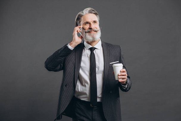 ビジネスとファッショナブルなひげそり成熟した男で灰色のスーツ電話で話す、コーヒーを飲むと灰色の背景に笑みを浮かべて. - 写真・画像