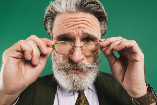 Vousatý učitel středního věku v khaki obleku drží brýle s rukama a mrkne očima se zábavou na zeleném pozadí - Fotografie, Obrázek