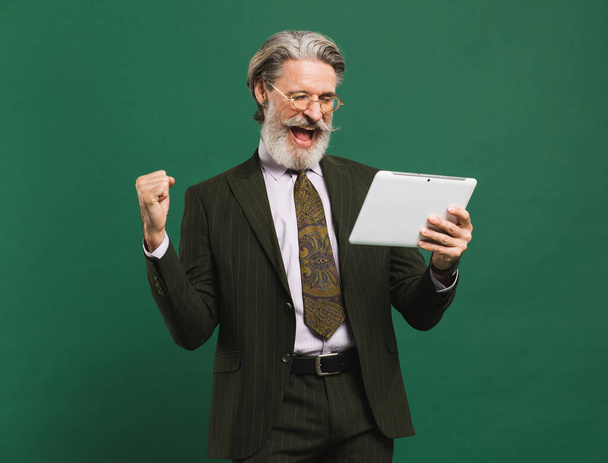 Szakállas középkorú férfi khaki öltönyben hangosan sikoltozik és táblagépre néz, miközben a boldogság öklét tartja a zöld háttér előtt. - Fotó, kép