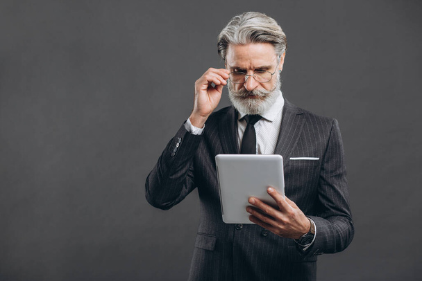 Homem maduro barbudo de negócios e moda em um terno cinza fazendo compras em tablet com seu cartão de crédito dourado no fundo cinza
. - Foto, Imagem
