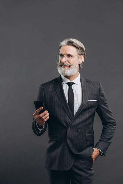 Üzleti és divatos szakállas érett férfi szürke öltönyben mosolyog és telefonon beszél a szürke háttér. - Fotó, kép