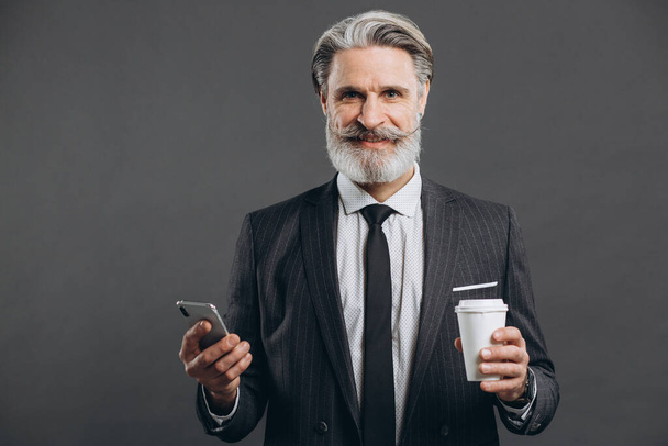 Obchod a módní vousatý dospělý muž v šedém obleku mluvit po telefonu, pít kávu a usmívat se na šedém pozadí. - Fotografie, Obrázek